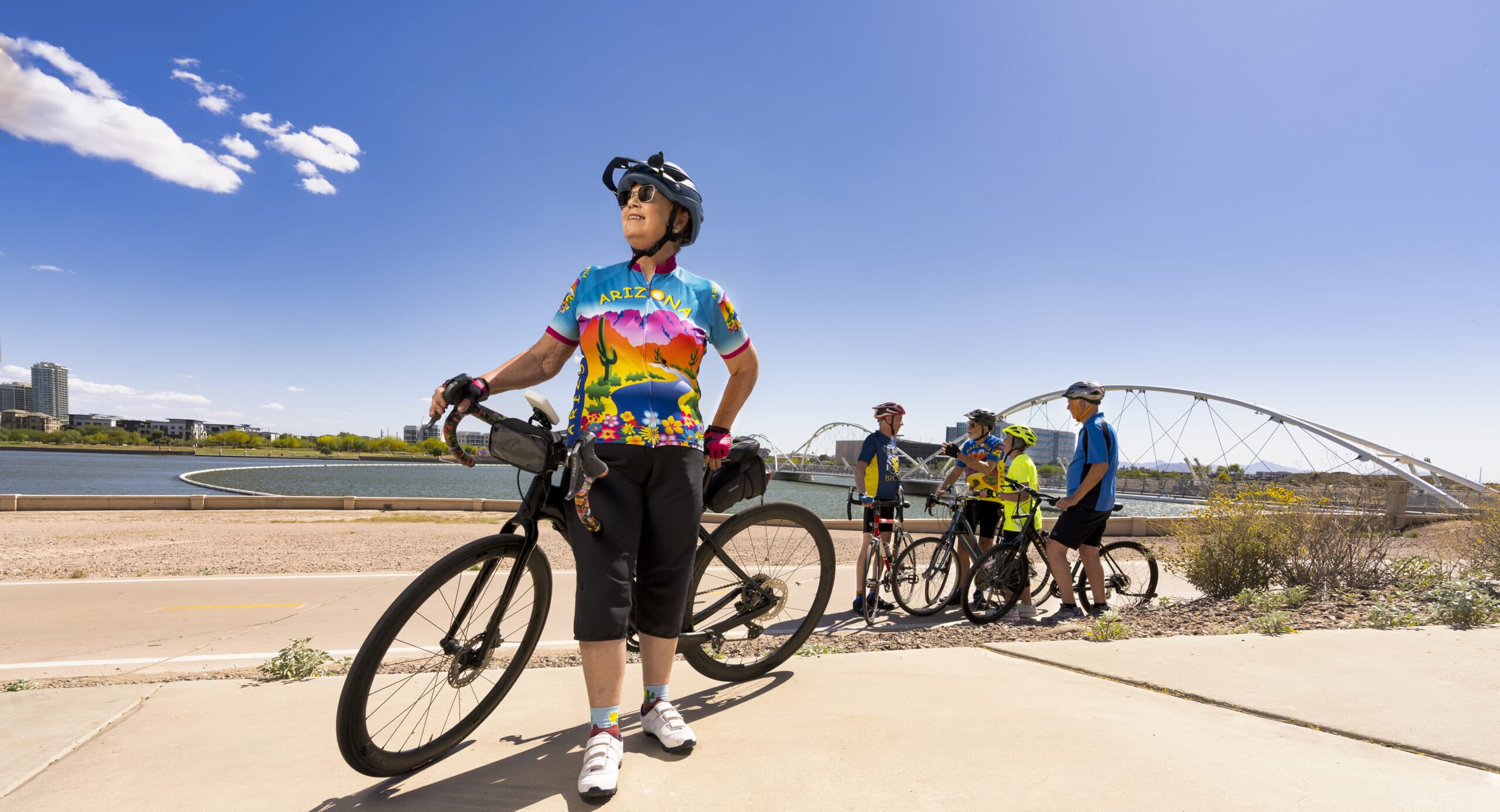 An older person stands with a bike in the foreground of the photo. A group of bikers stands in front of a picturesque lake in the background.